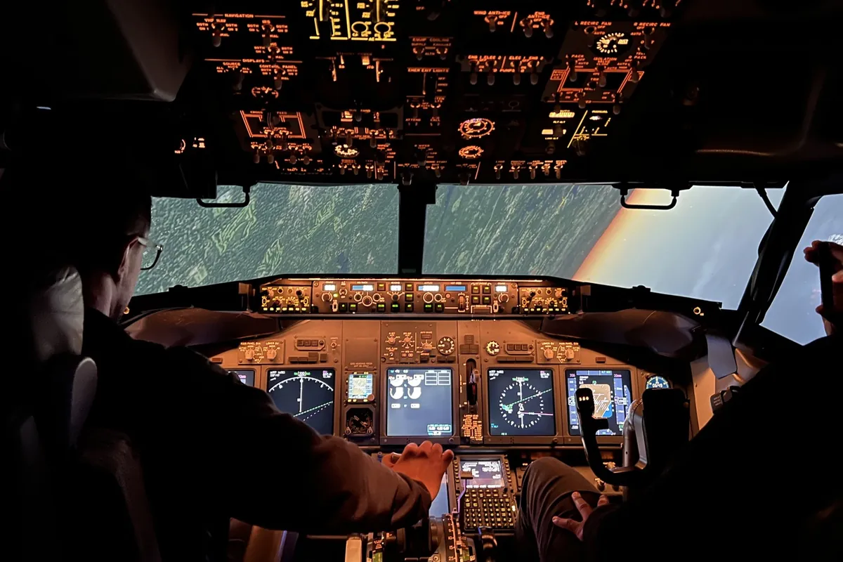 Two pilots flying a Boeing 737 full flight simulator, seen from behind, with the overhead panel and flight displays lit