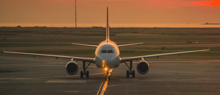 Airbus A320 jet taxiing head-on along a lit taxiway centerline at sunset, under an orange sky with water on the horizon