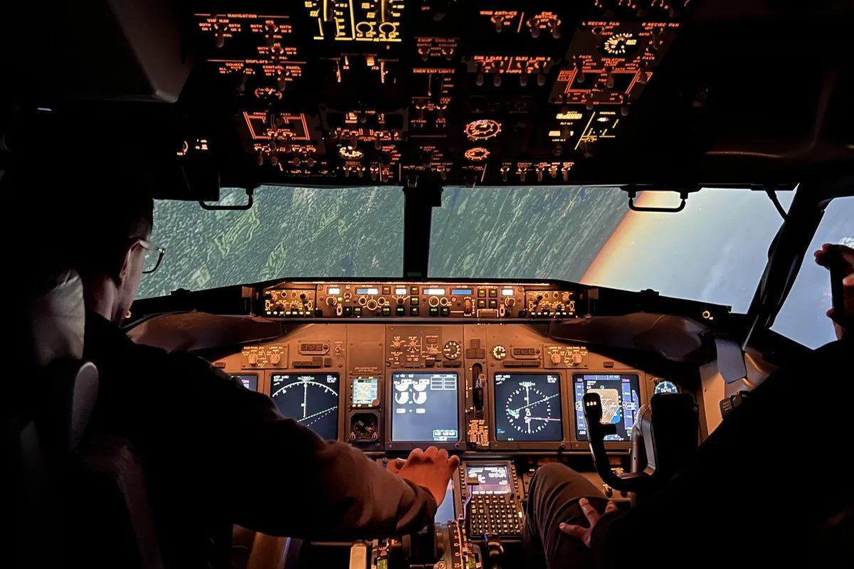 Two pilots flying a Boeing 737 full flight simulator, seen from behind, with the overhead panel and flight displays lit